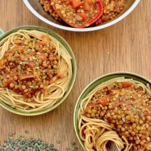Two bowls of spghettis topped with lentil ragu next to uncooked green lentils on a board.