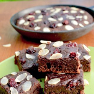 Squares of chocolate brownie topped with cranberries and almonds, in front of a skillet