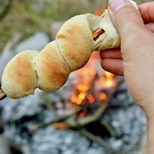 A hand tears open a bread on a stick cooked on a campfire