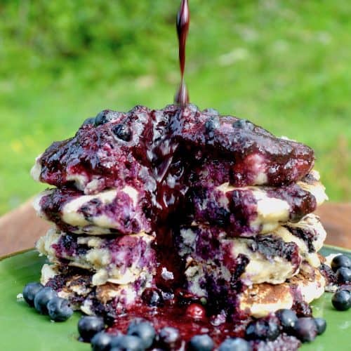 Pouring a berry sauce over a stack of vegan pancakes