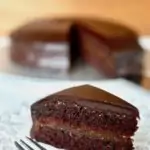 A slice of sachertorte on a plate, with the remaing chocolate cake behind
