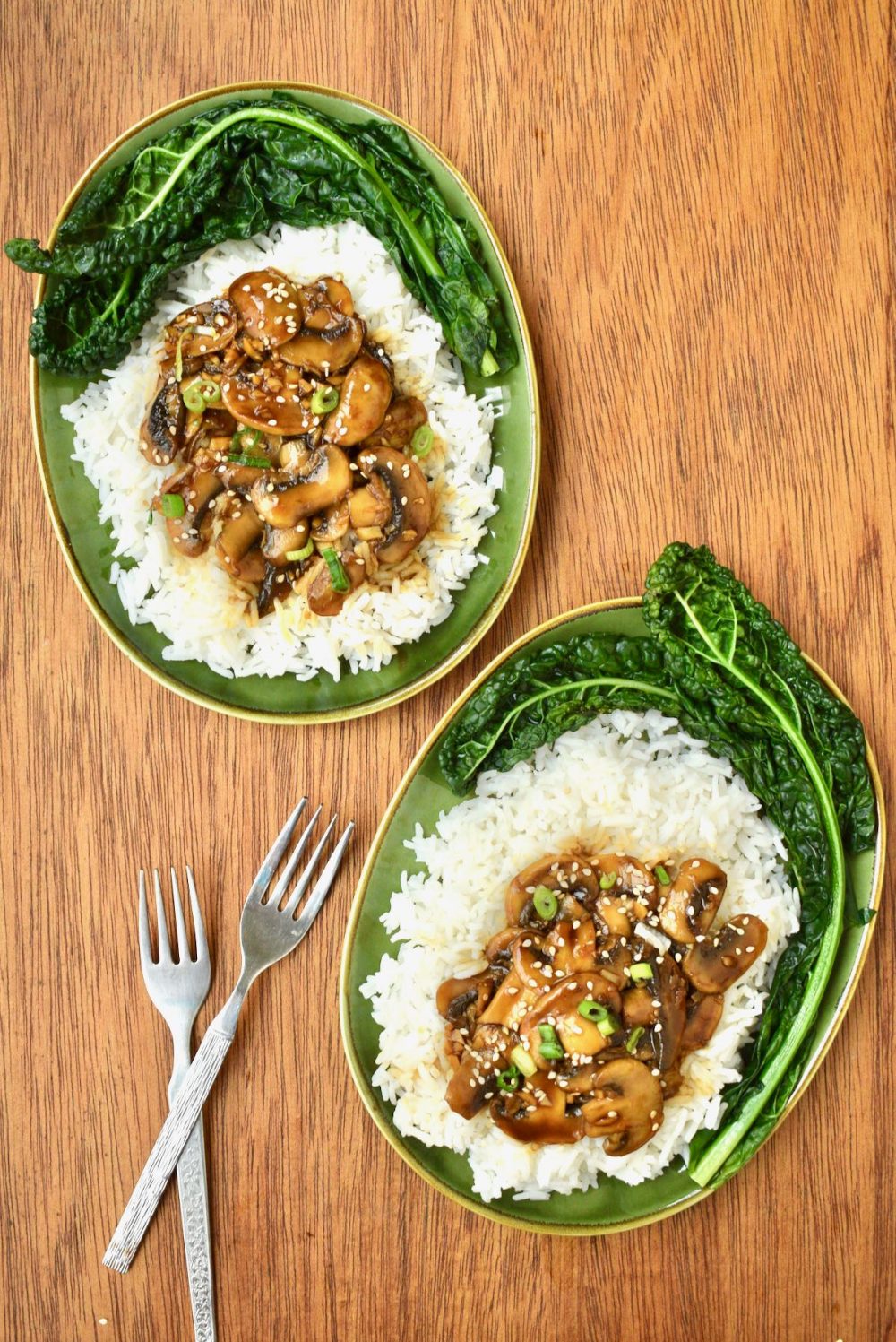 Two green plates with rice, steamed kale and teriyaki mushrooms garnished with sesame seeds and spring onion