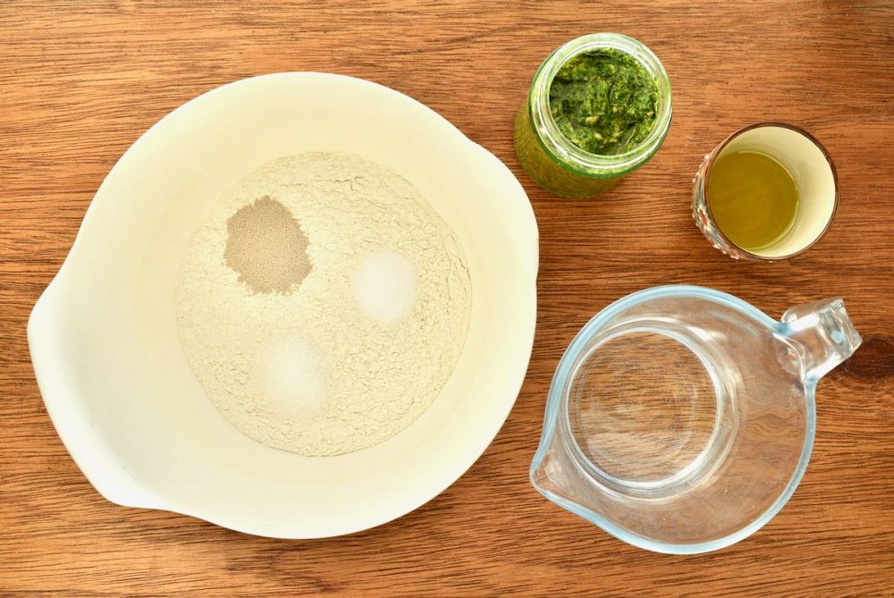 Ingredients for the bread on a wooden board - a bowl with flour, yeast, sale and sugar, a jug of water, a jar of pesto, and some olive oil in a dish