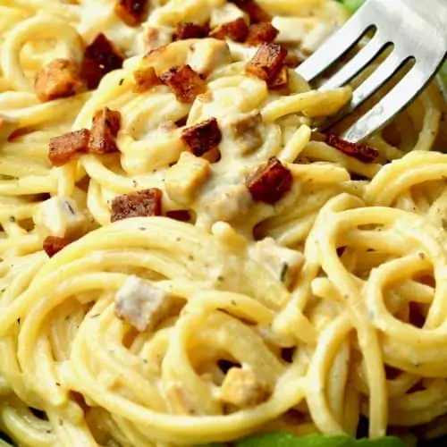 A fork digs in to a plate of creamy spaghetti carbonara with smoked tofu bits on top