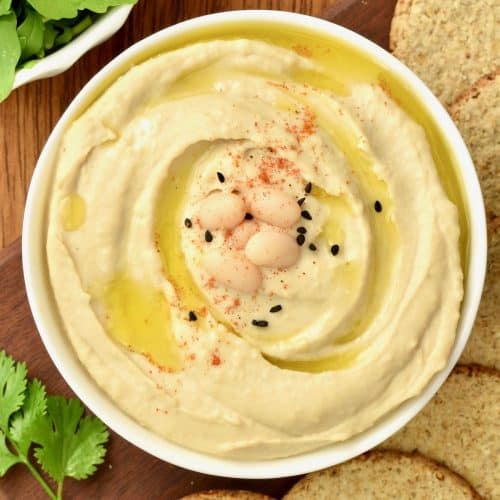 A white bowl of creamy hummus garnished with cannellini beans, paprika and nigella seeds
