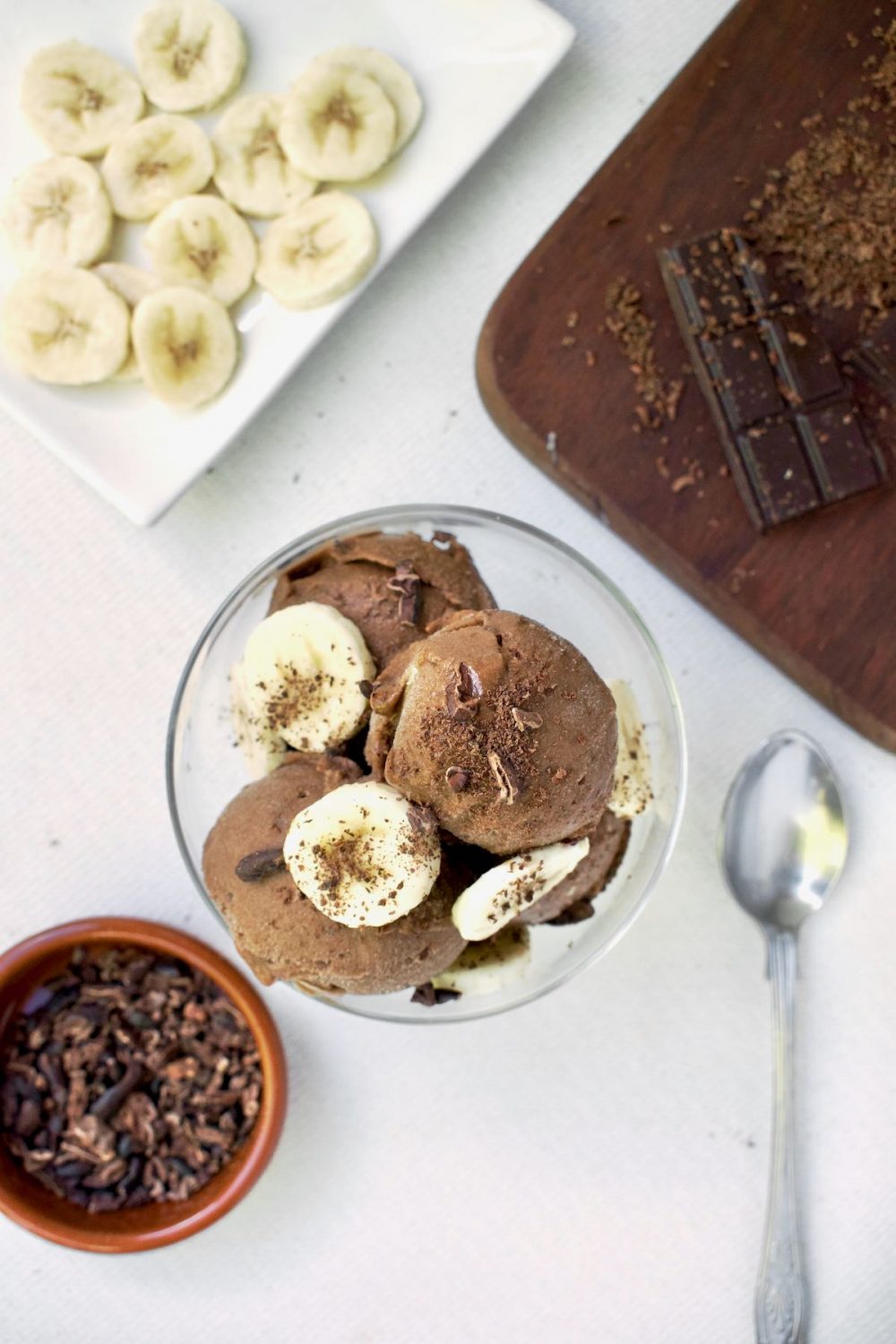 A bowl of nice cream seen from above next to a dish of cocoa nibs, banana slices and grated chocolate