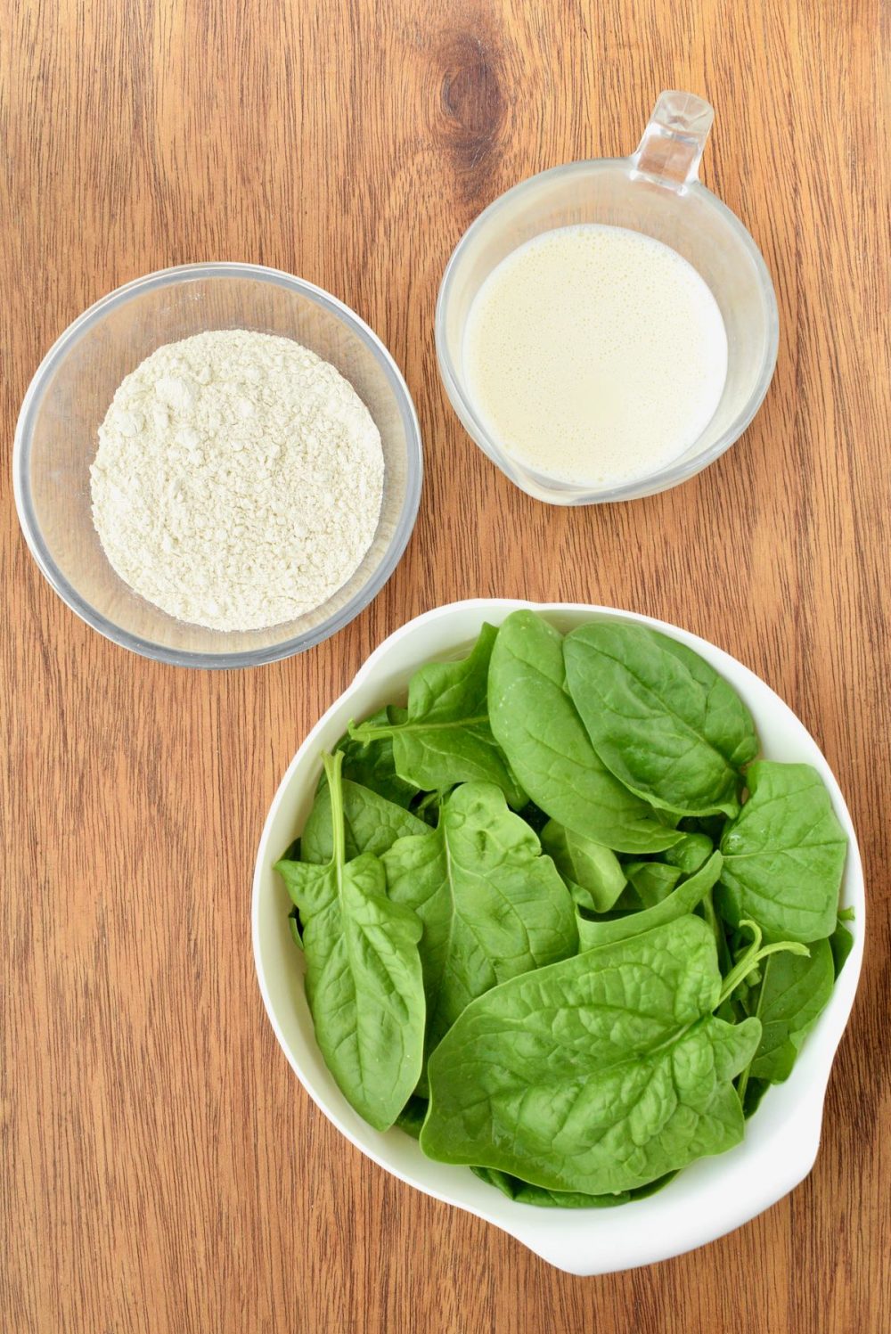 A bowl of flour, jug of soy milk and a bowl of spinach on a wooden board 