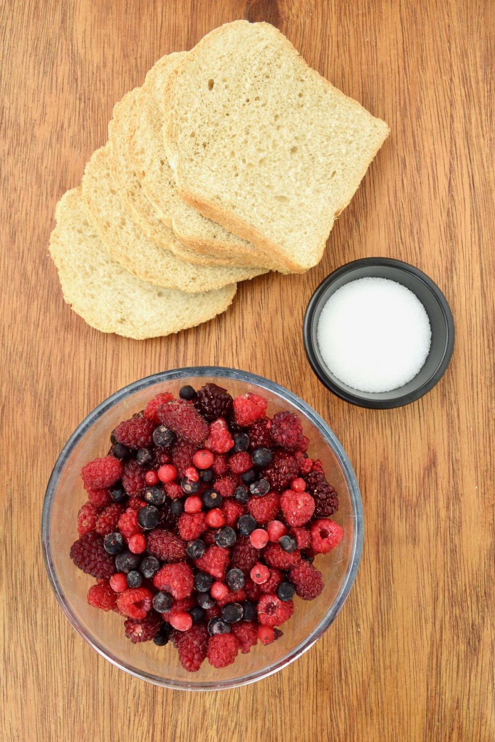 The ingredients for the summer pudding laid out on the table: slices of white bread, a small bowl of sugar and a big glass bowl filled with a variation of red berries.