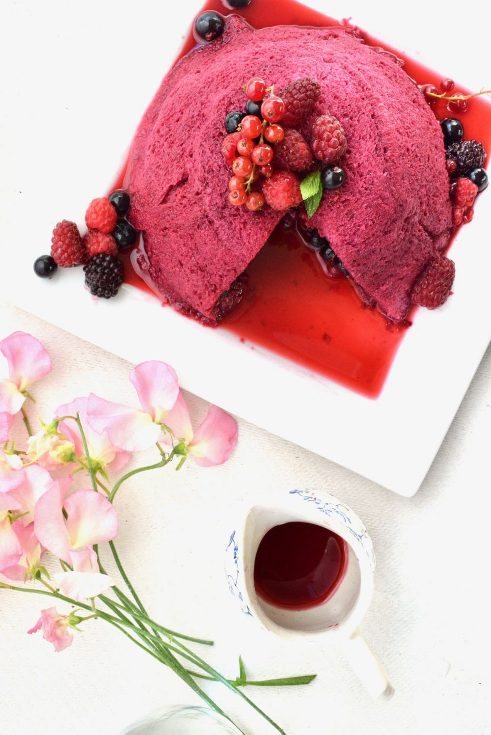 A top down view of the decorated pudding on a square white plate. Next to it there's a small jug of dark red berry juice while a bunch of delicate pink sweet pea flowers add some atmosphere. A big cake slice has been cut from the pudding and the juice fills the plate all around it.