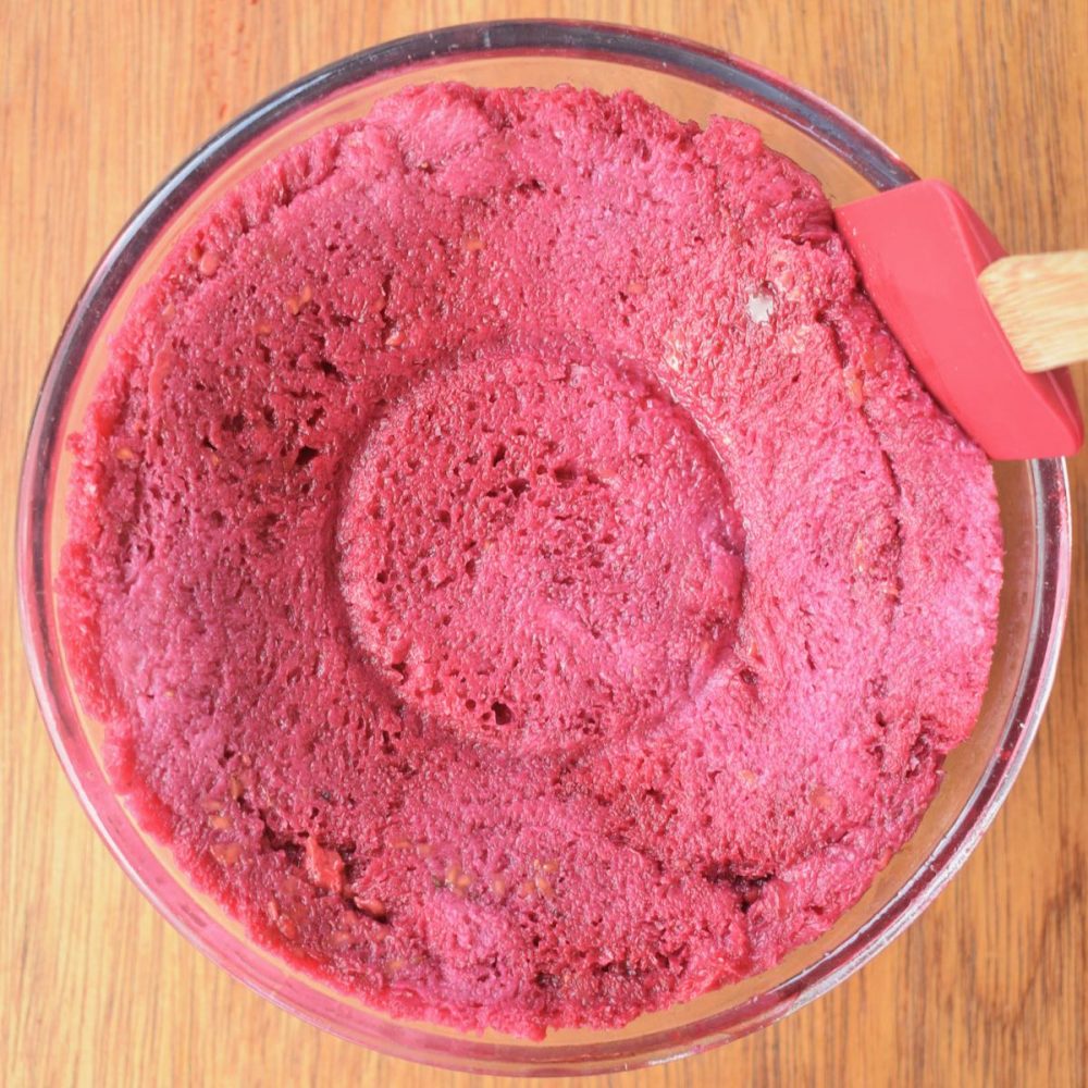 A red silicon spatula is moved around the side of the pudding inside the glass bowl. Still visible is a ring imprinted on the top of the pudding where the plate has been sitting.