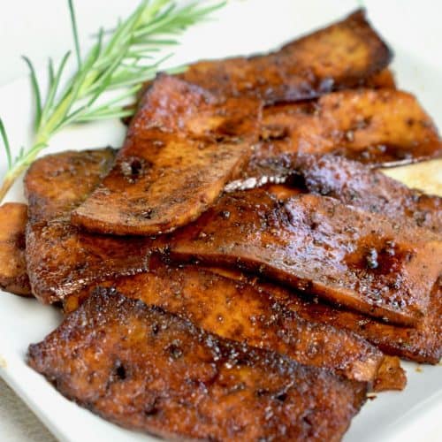 A juicy pile of dark brown, fried slices of tofu on a white square plate, decorated with a twig of fresh rosemary.
