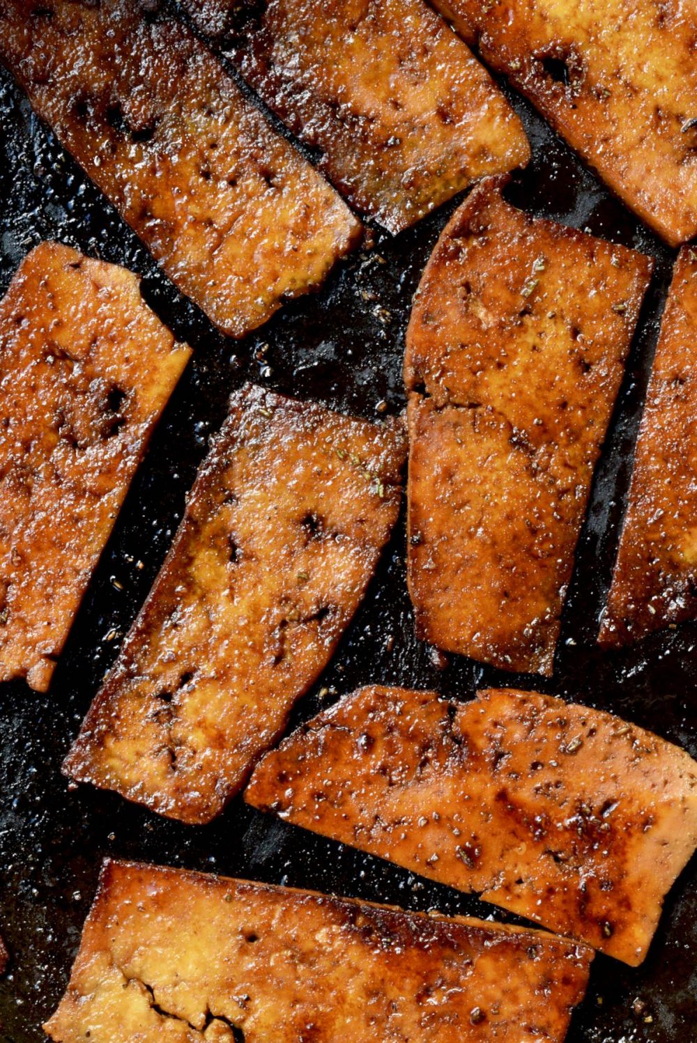 A top-down close-up of several deep brown fried slices of marinated tofu in a frying pan.
