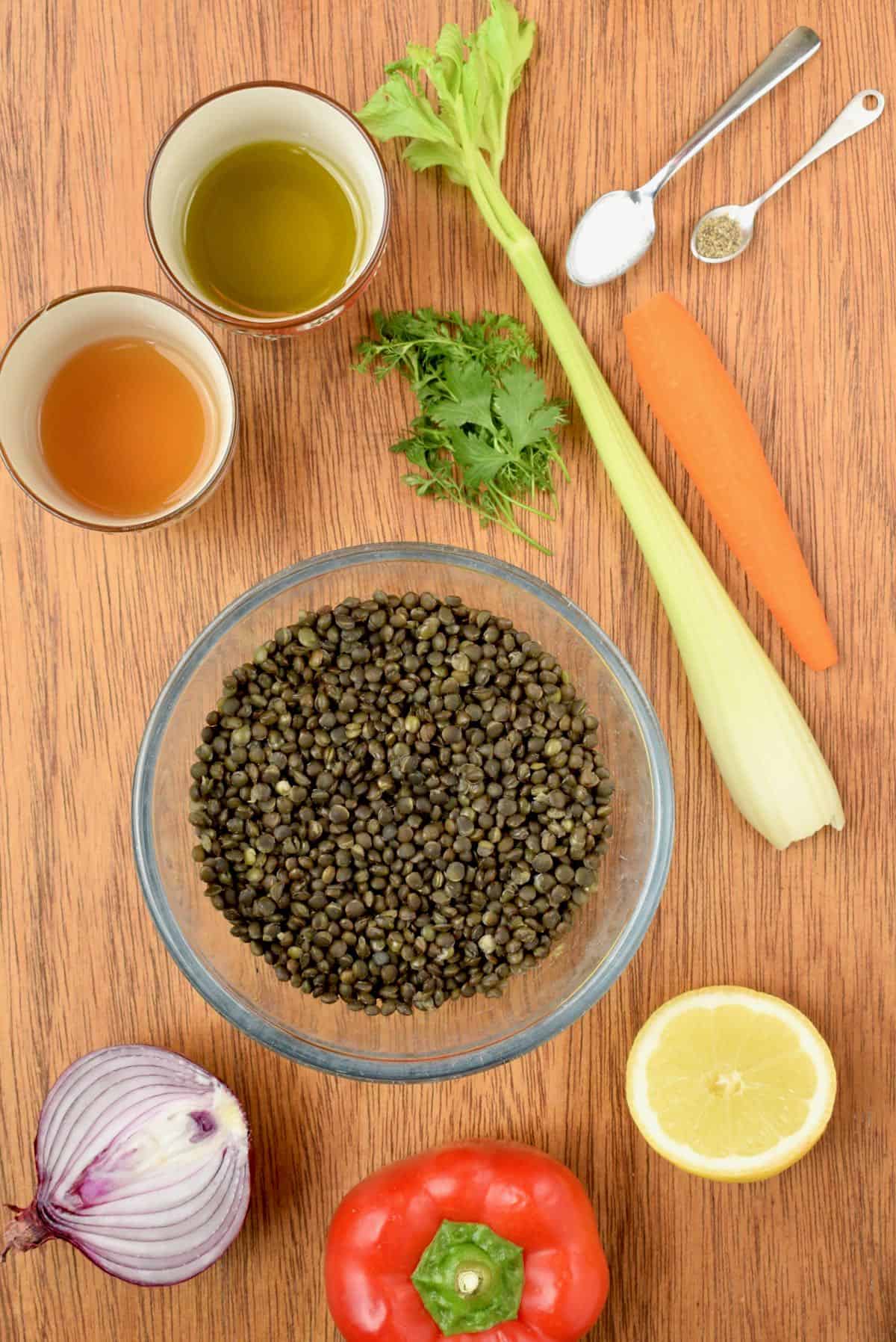 All the ingredients on a wooden board, a bowl of cooked lentils, raw vegetables, salt, pepper, half a lemon and two pots of oil and vinegar. 