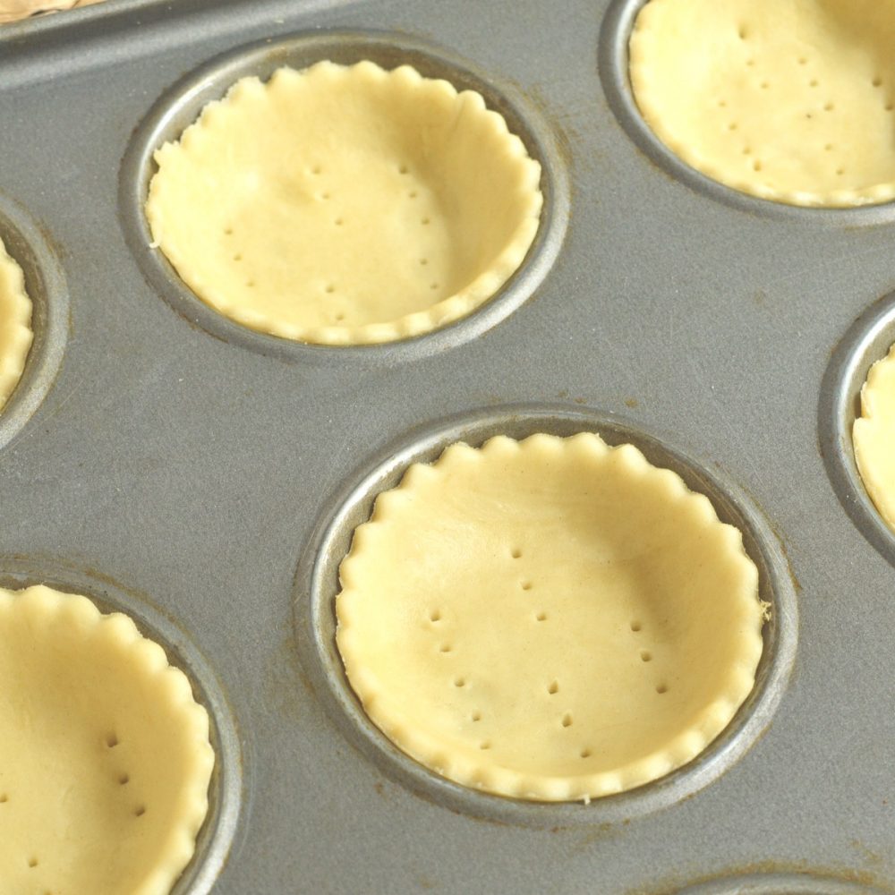 Small rounds of pastry in a shallow muffin tin are pricked several times with a fork and ready to be blind baked.