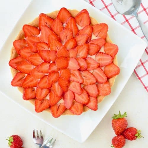 A bright red strawberrry tart served on a square plate on a white table. Next to it a cake server on a white and red tablecloth, some strawberries and two forks.