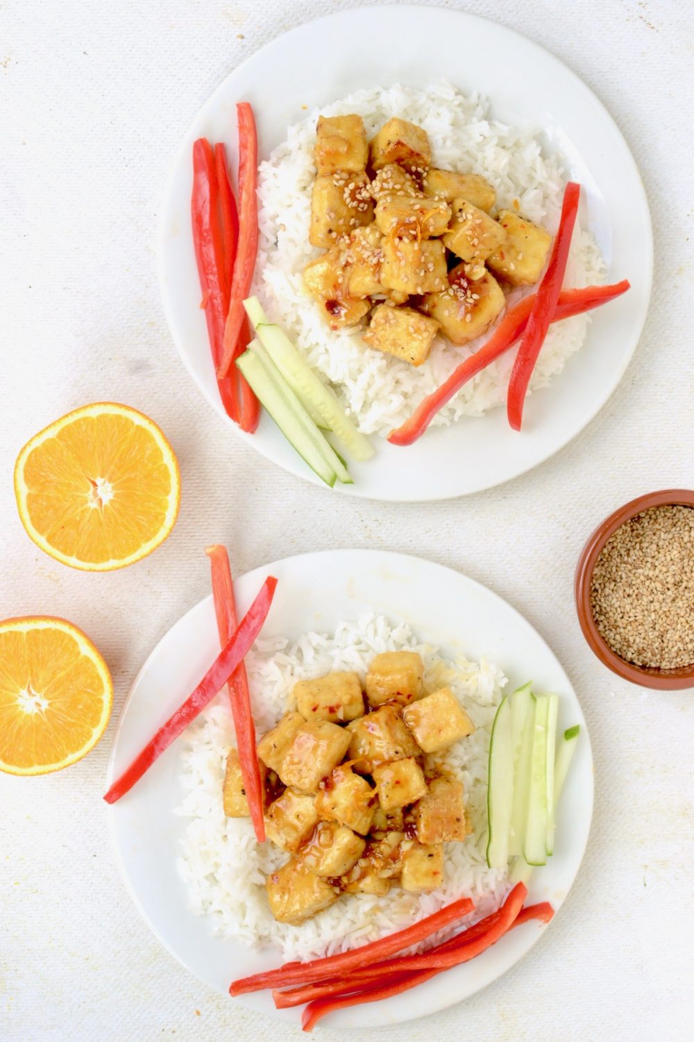 Two servings of Orange Tofu with white rice and raw vegetables, next to two orange halves and a small pot of toasted sesame seeds viewed from above.