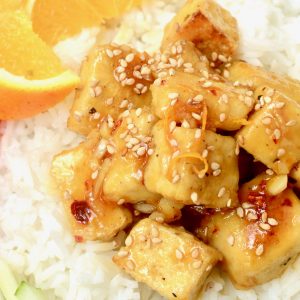 A close up shot of orange tofu sprinkled with toasted sesame seeds, served on white rice and with a wedge of fresh orange.