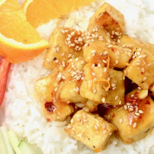 A close up shot of orange tofu sprinkled with toasted sesame seeds, served on white rice and with a wedge of fresh orange and raw vegetables.