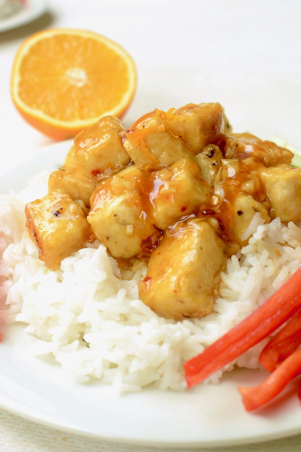 A portion of orange tofu, coated thickly in sauce and served with some white rice alongside some fresh vegetables. A half orange in the background.