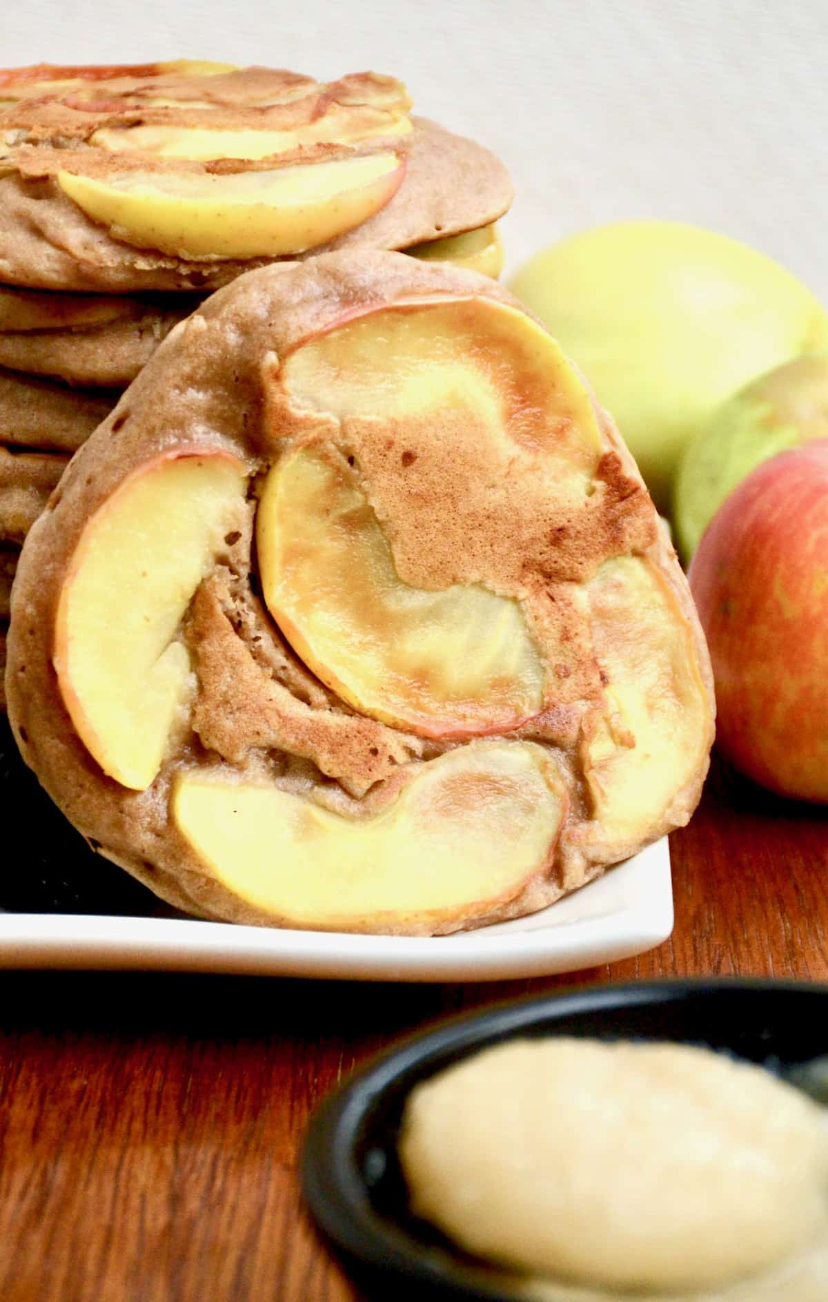 A pancake topped with apple slices is leaned against pancake stack.