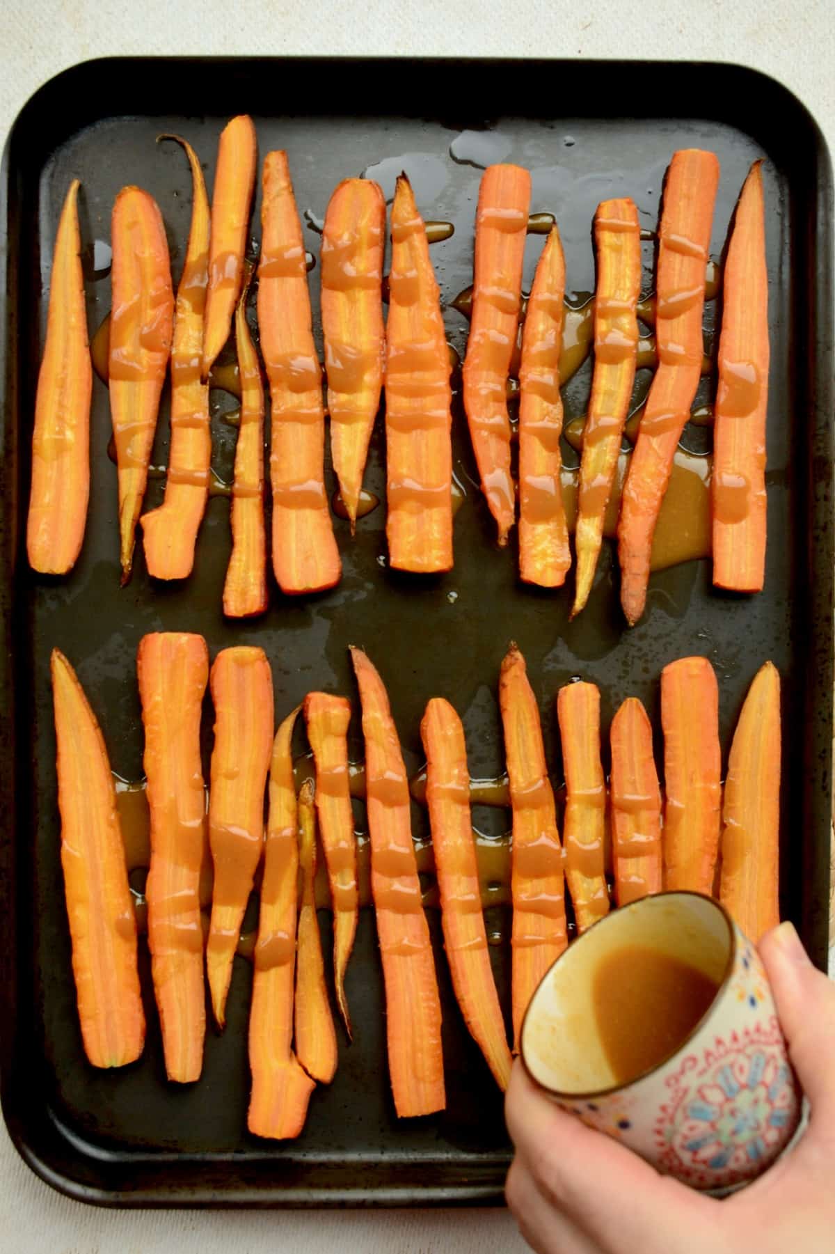 A brown viscous glaze is drizzled onto carrots on a roasting tray.