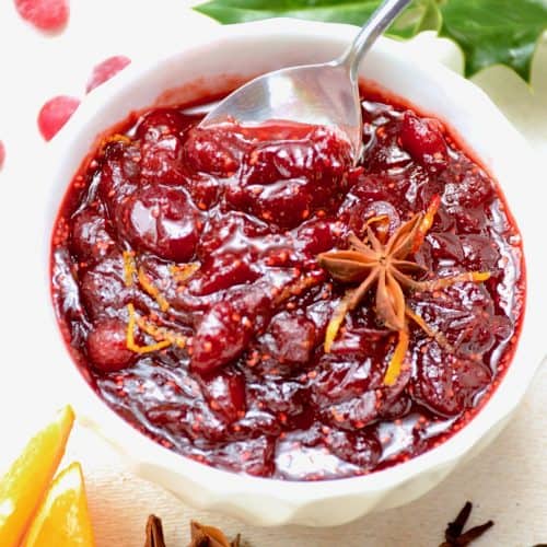 A white bowl of dark red cranberry sauce, garnished with orange zest and star anise.