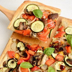 A corner of the vegetable tart has been cut and is being picked up by a wooden spatula.
