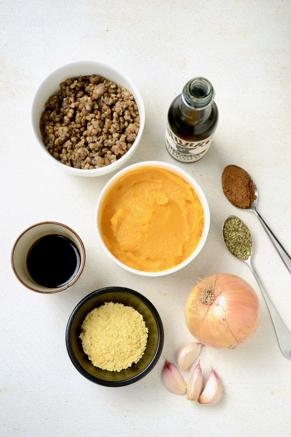 Ingredients on a white surface, bowls of pumpkin puree, lentils, nutritional yeast, soy sauce, a bottle of liquid smoke, onion, garlic and spices on spoons.