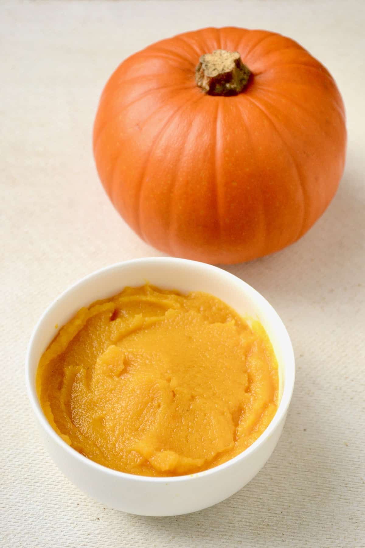 Orange coloured homemade pumpkin puree in a bowl next to a small pumpkin.