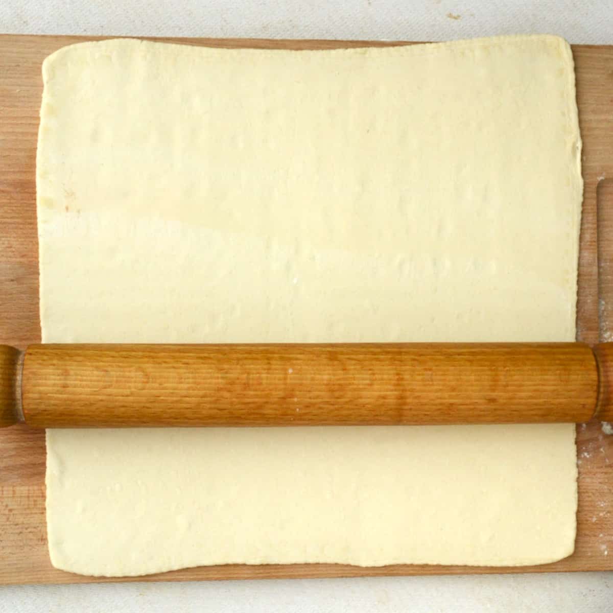 Puf pastry is rolled out on a wooden board using a rolling pin.