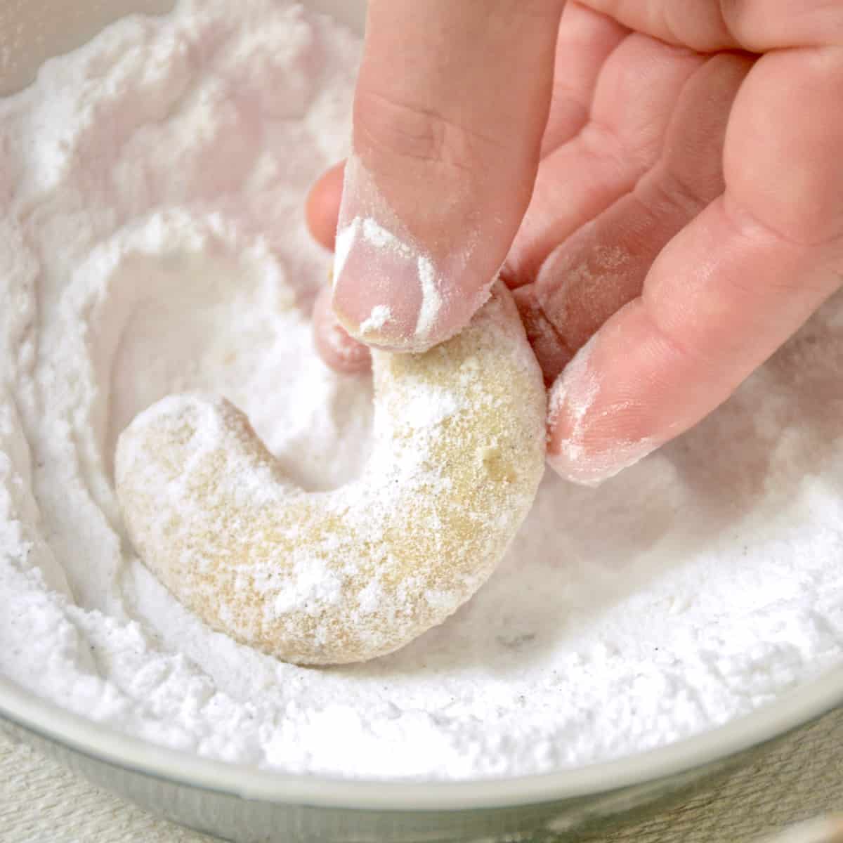 A crescent cookie is dipped into a bowl of powdered vanilla sugar to completely coat it.