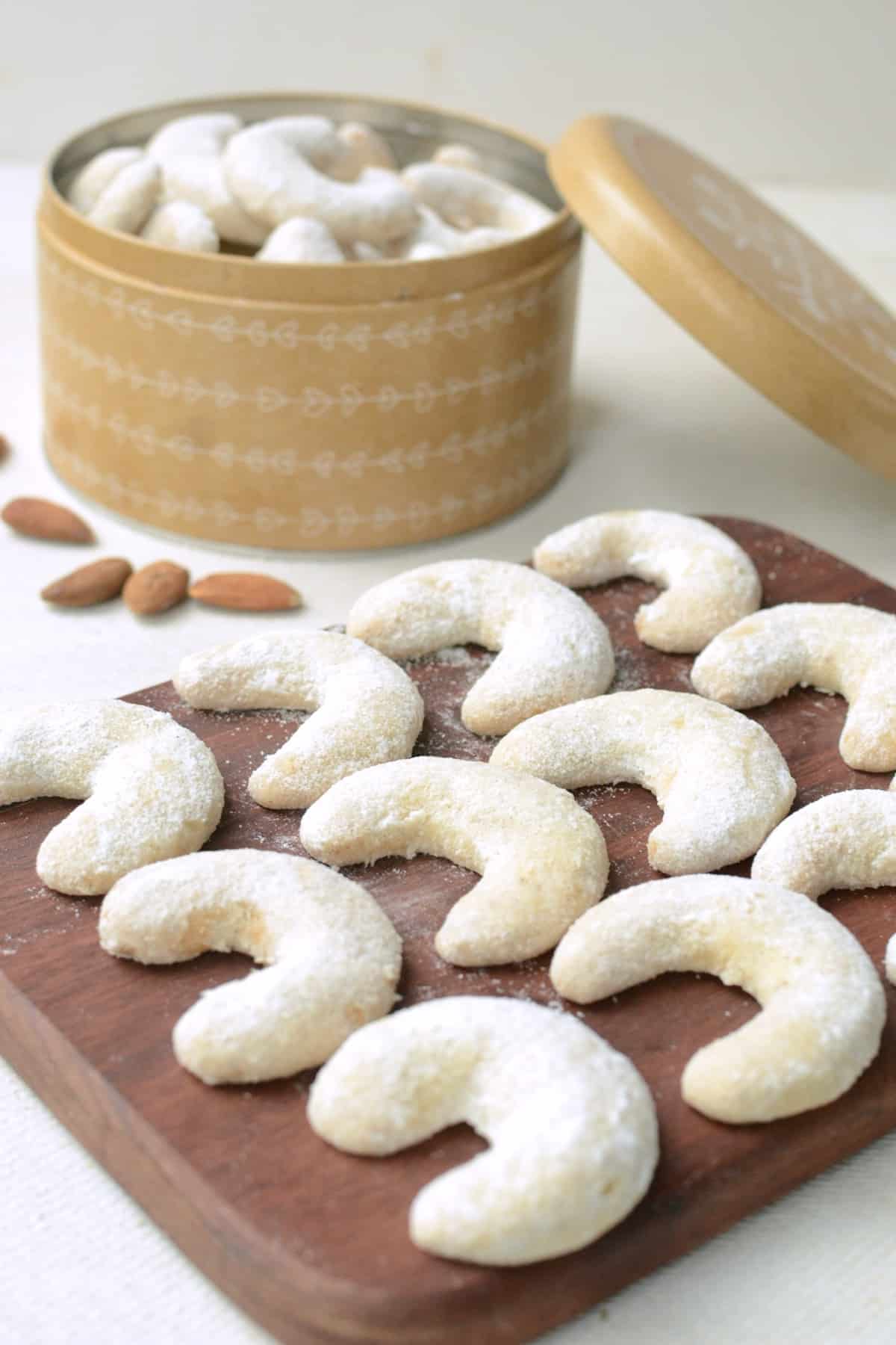 Rows of crescent shaped, sugar dusted cookies lined up on a dark brown wooden board. In the background, a light brown, round tin full of cookies.