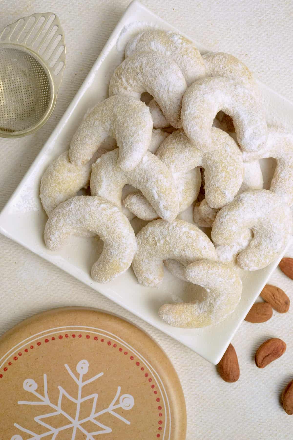 A white square plate fully loaded with crescent-shaped, sugar dusted cookies.