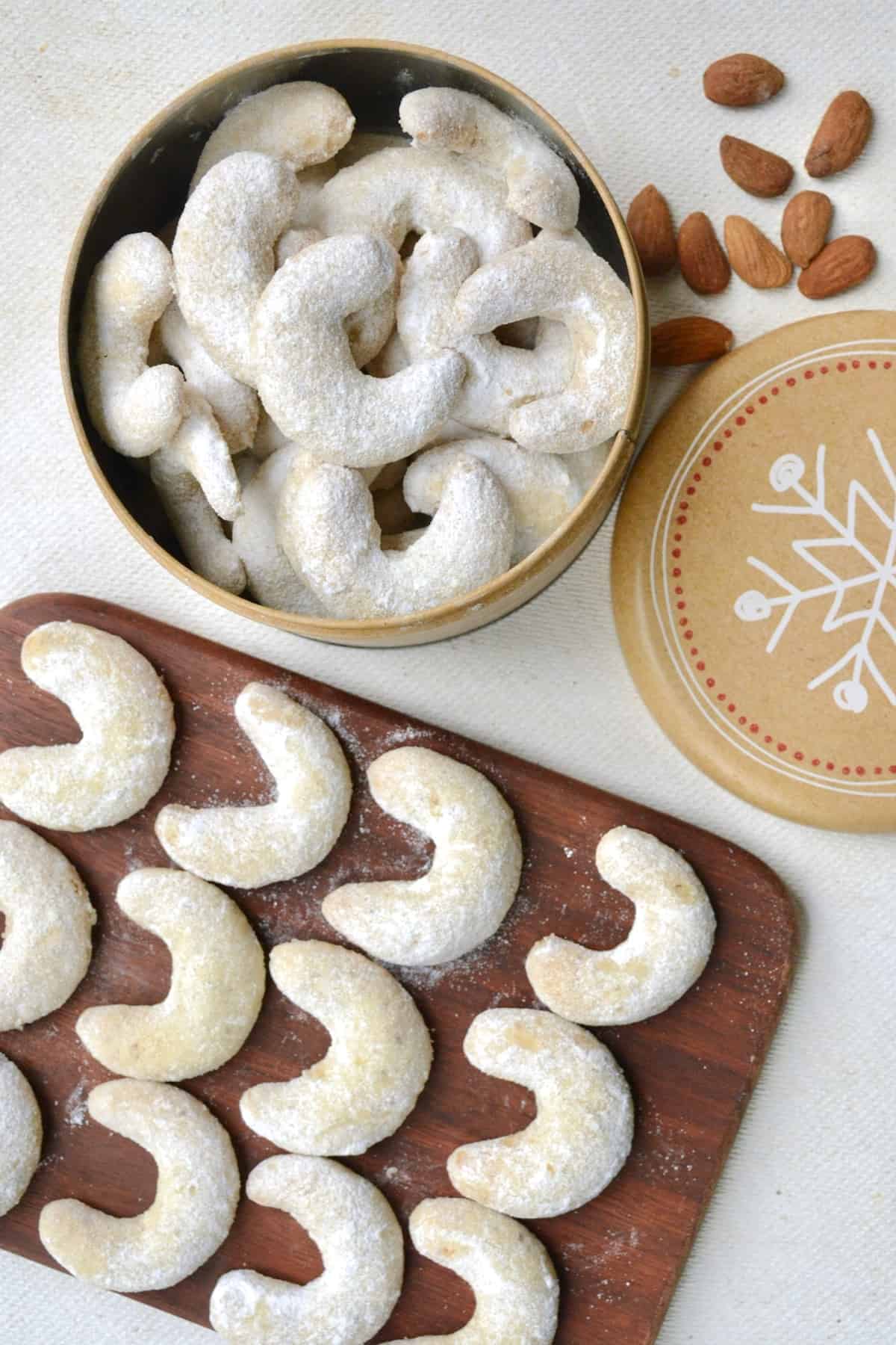 Sugar dusted crescent cookies lined up in rows of four on a dark wooden board, next to a round tin full of more cookies.