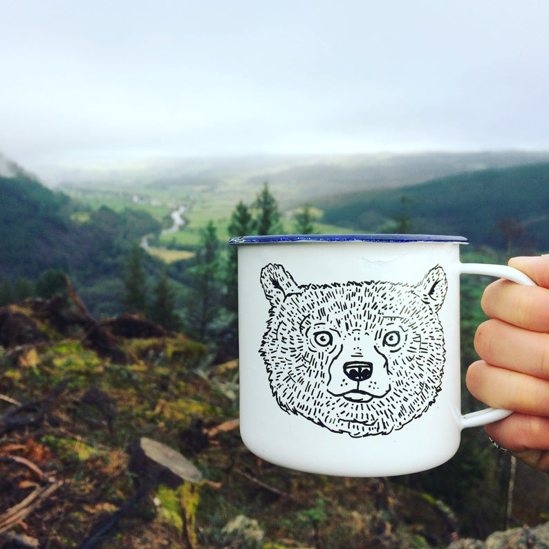 A hand holds up a white enamel mug with a bear drawing on it, in front of a forest landscape backdrop.