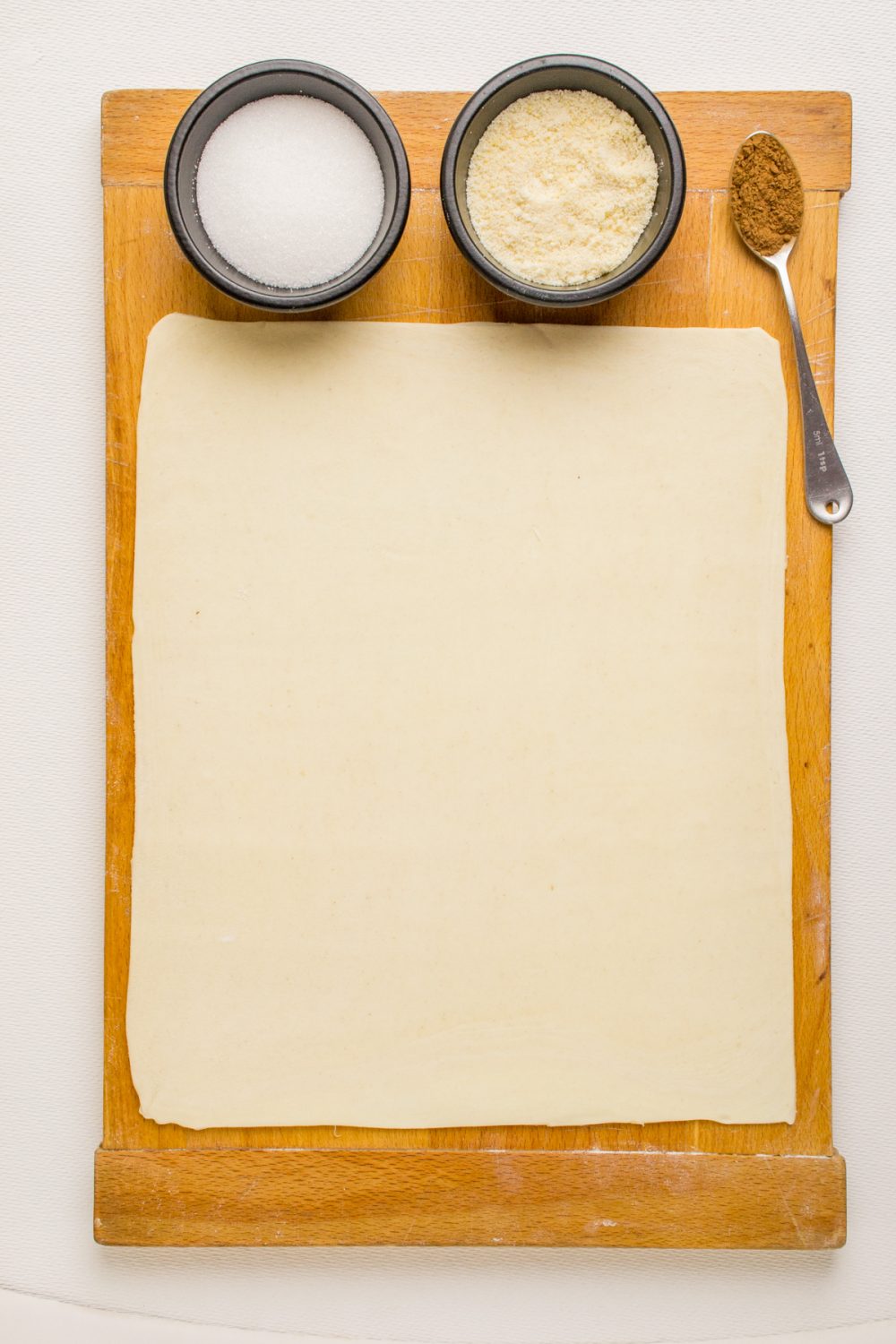 A sheet of puff pastry, small bowls of sugar and ground almonds and a spoonful of cinnamon on a wooden board.