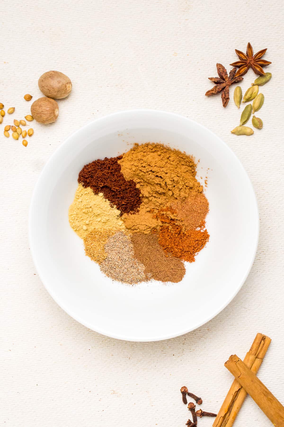 A white bowl of ground spices, whole spices on the surface around the bowl.