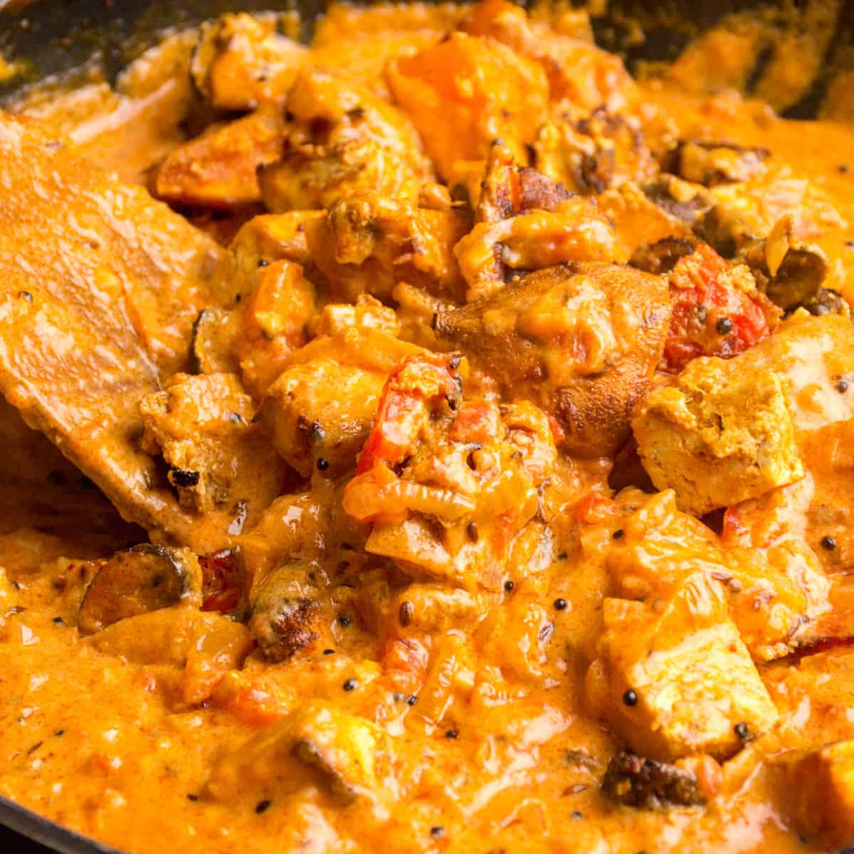 A wooden spatula stirs in the vegetables and tofu tikka to the masala sauce.
