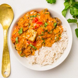 A bowl of richly coloured vegan tikka masala with rice, a brass spoon and herbs on the next to it..