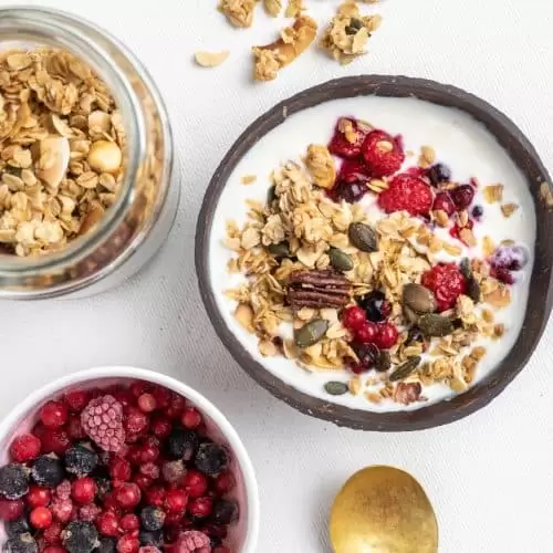 A bowl of granola with yoghurt and berries next to a jar of granola and a bowl of fruit.