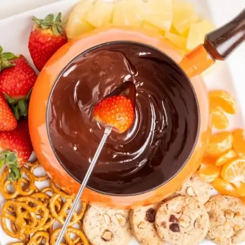 Dipping a strawberry into a chocolate fondue in a pan, surrounded by cookies and fruit.