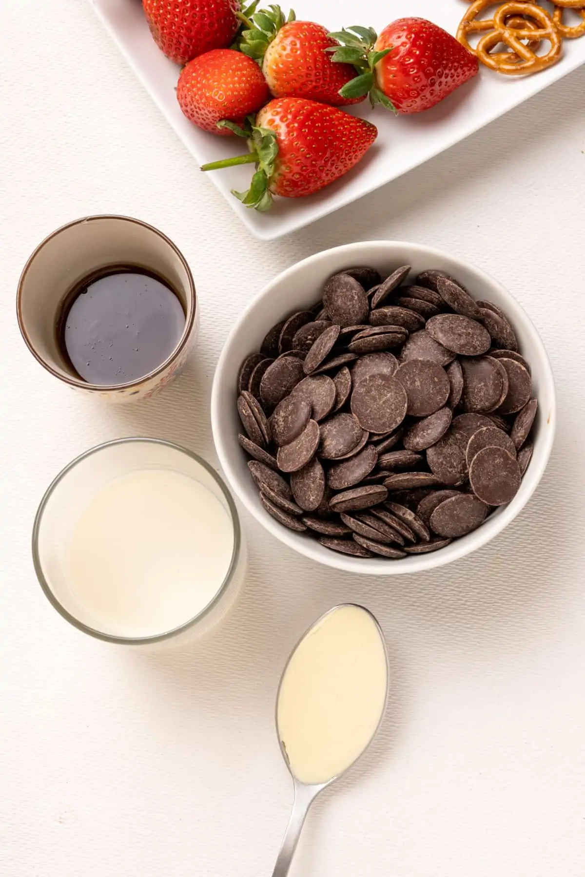 A bowl of chocolate buttons, a glass of oat milk, spoonful of vegan butter, a dish of maple syrup and some strawberries on a plate.