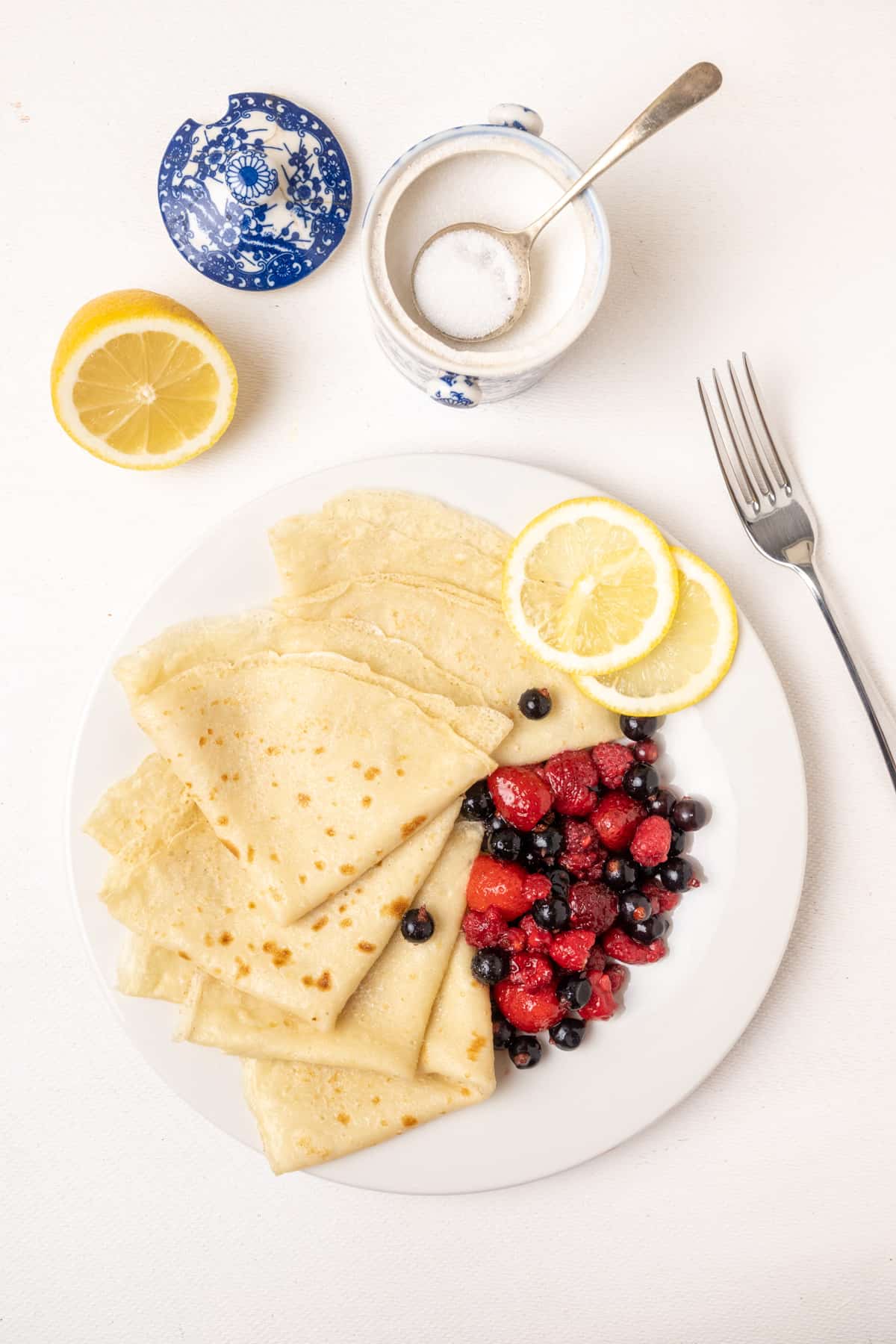 A plate of thin vegan pancakes with lemon and a sugar bowl next to it.