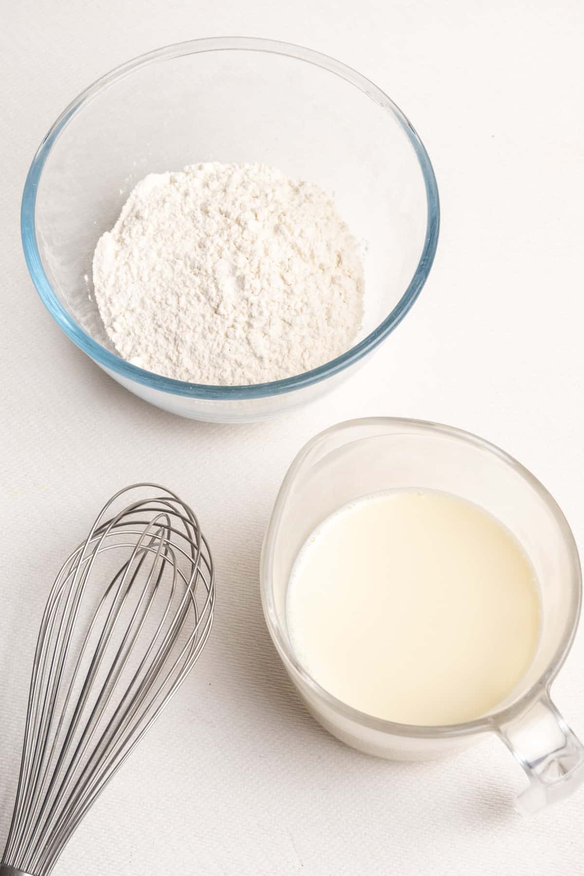A bowl of flour, a jug of soy milk and a whisk on a white background.