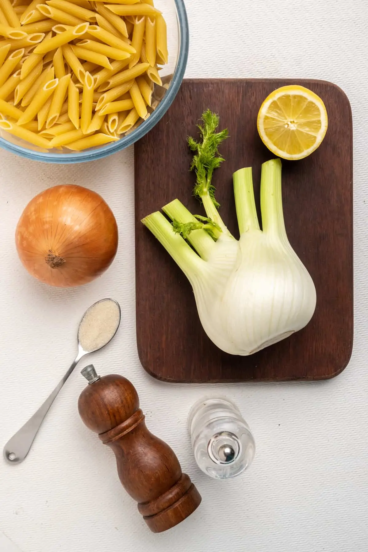 The ingredients for the fennel pasta laid out on a table: Penne style pasta, a bulb of fennel, a large onion, a lemon for the lemon juice, and some sugar, salt and pepper.