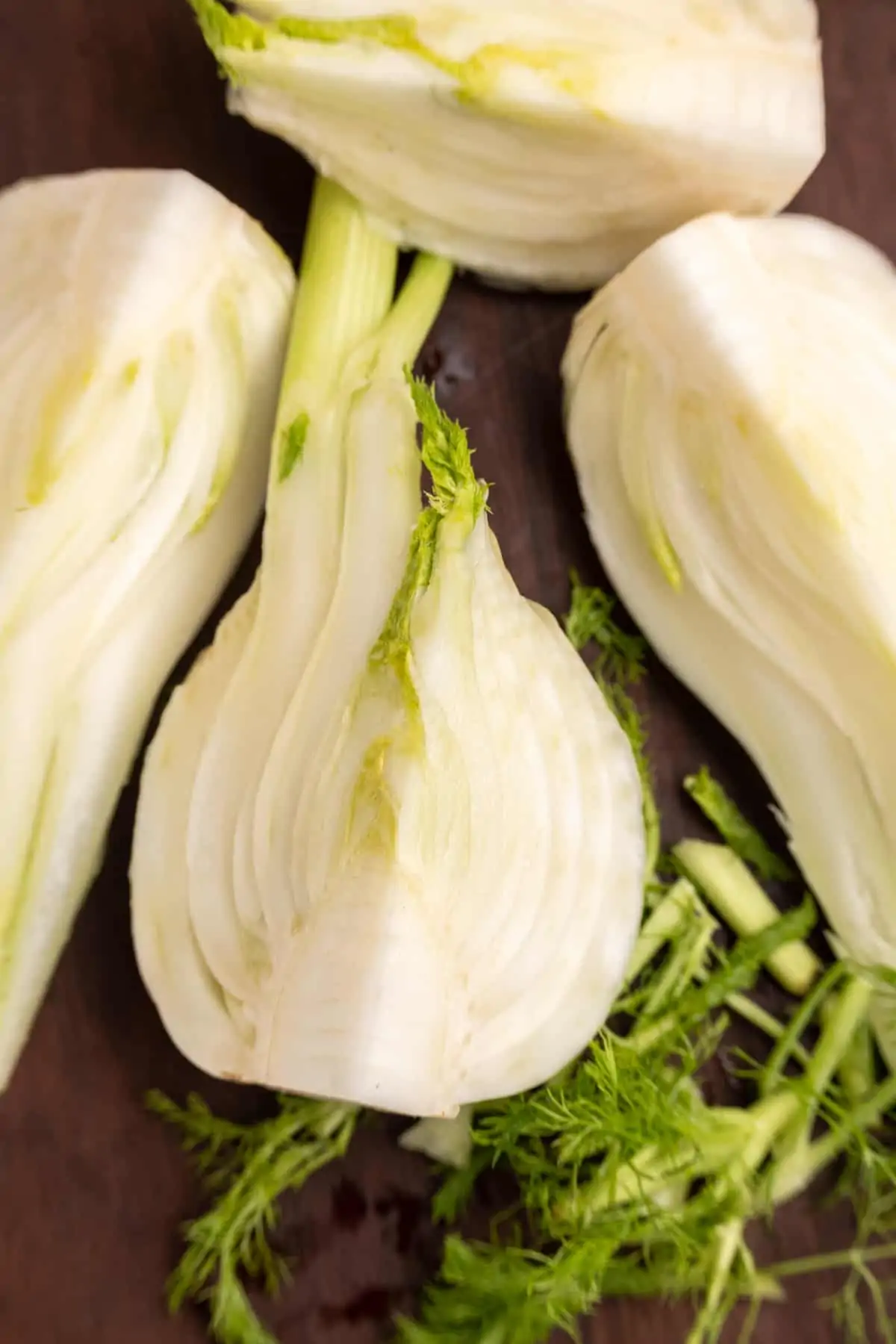 A bulb of fennel cut into quarters. The quarter bulbs are sitting on a wooden board, and the green fronds have been chopped off and set aside.