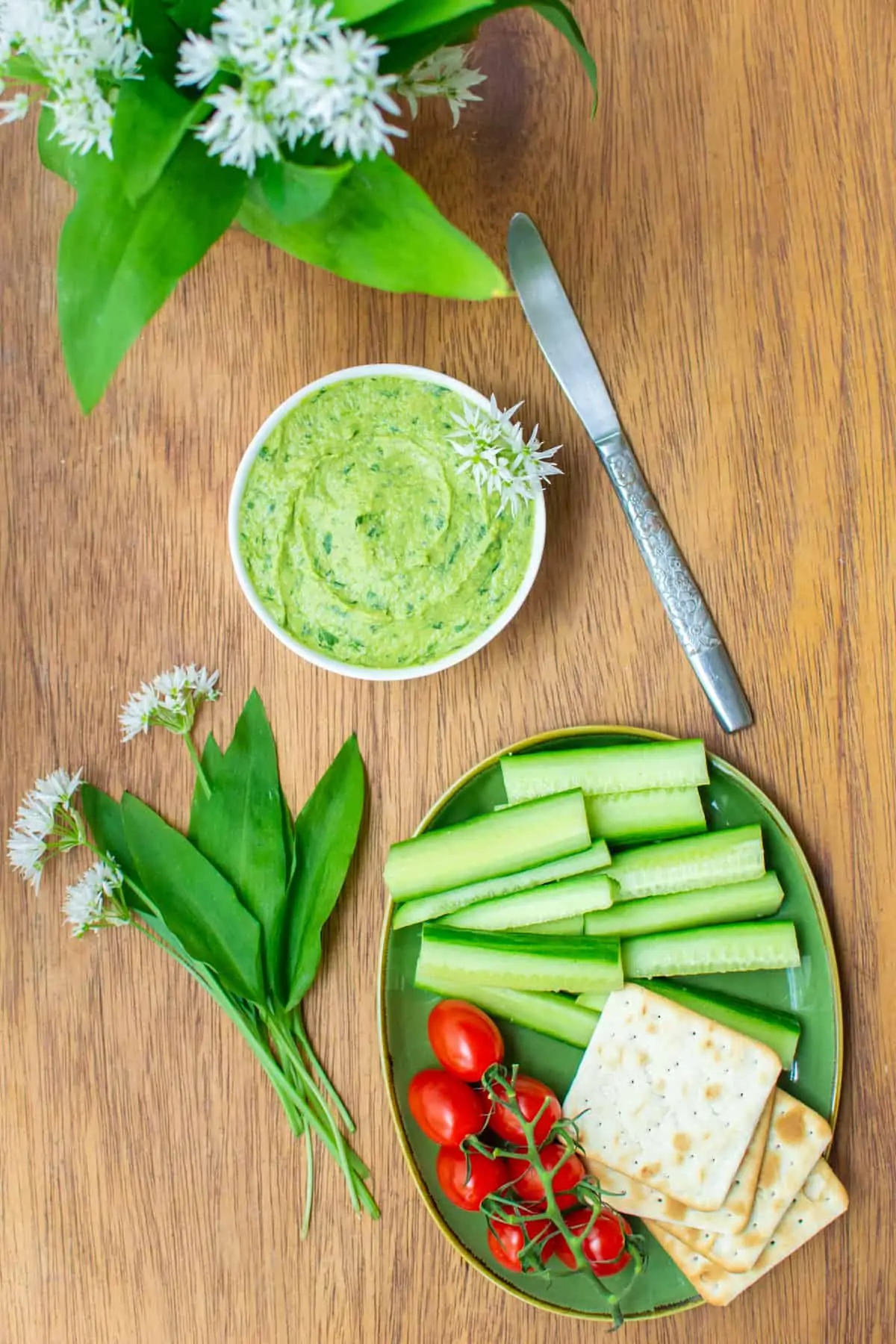A bowl of green hummus decorated with wild garlic flowers, next to a plate of crudités and crackers.