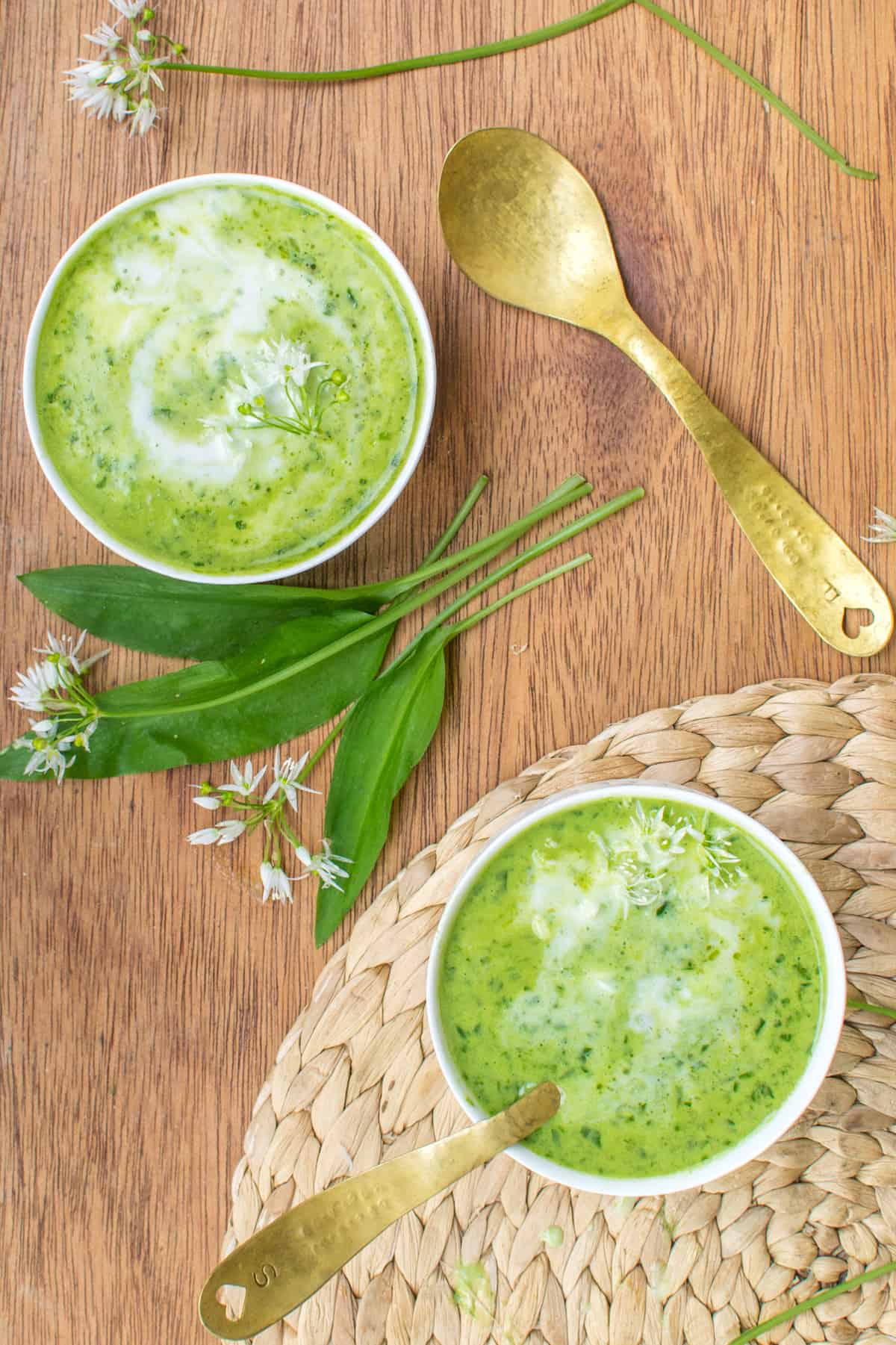 Two bowls of green soup, one of them with a spoon in it. Between the bowls some fresh wild garlic leaves and flowers.