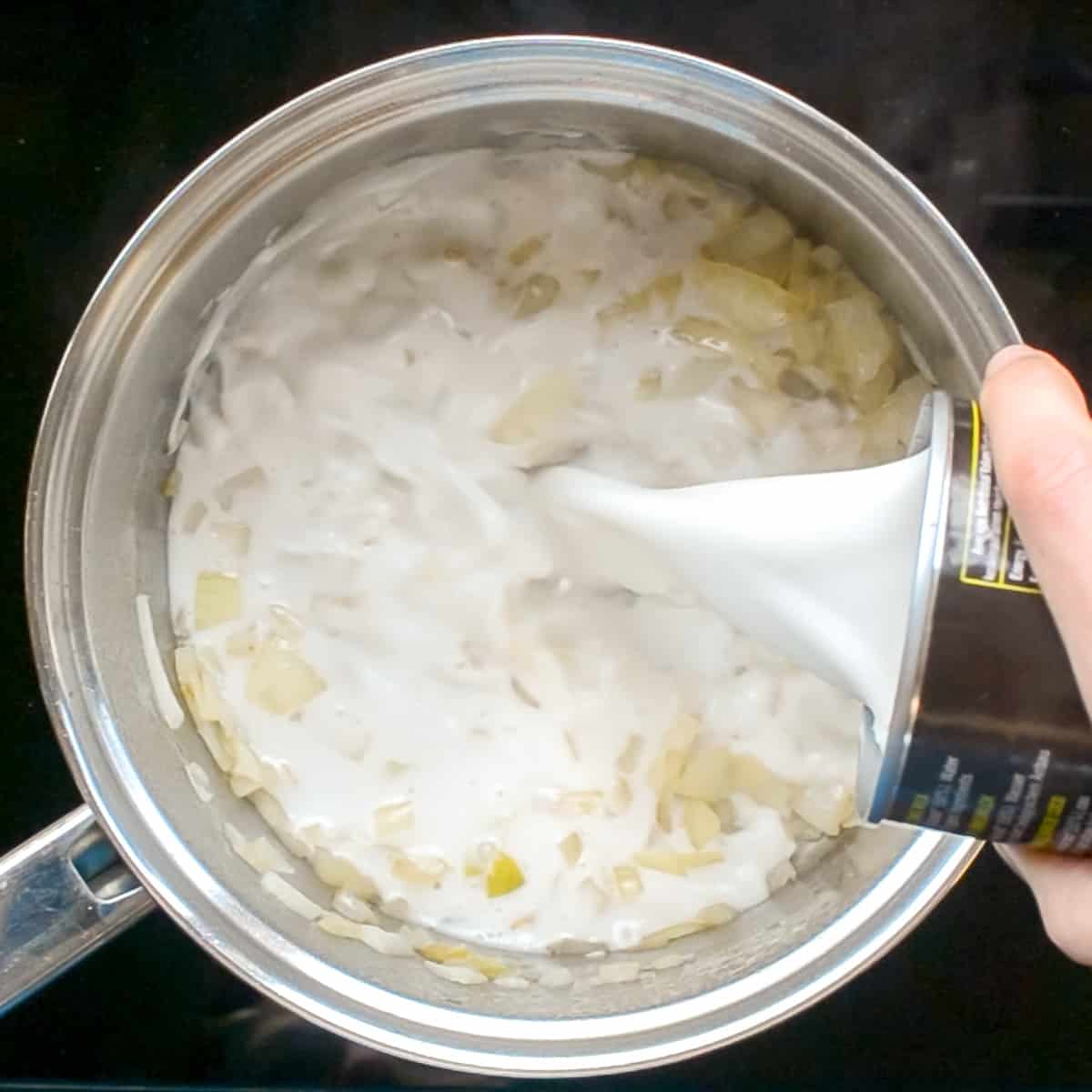 Coconut milk is poured from a can into the saucepan with sauteed onions.