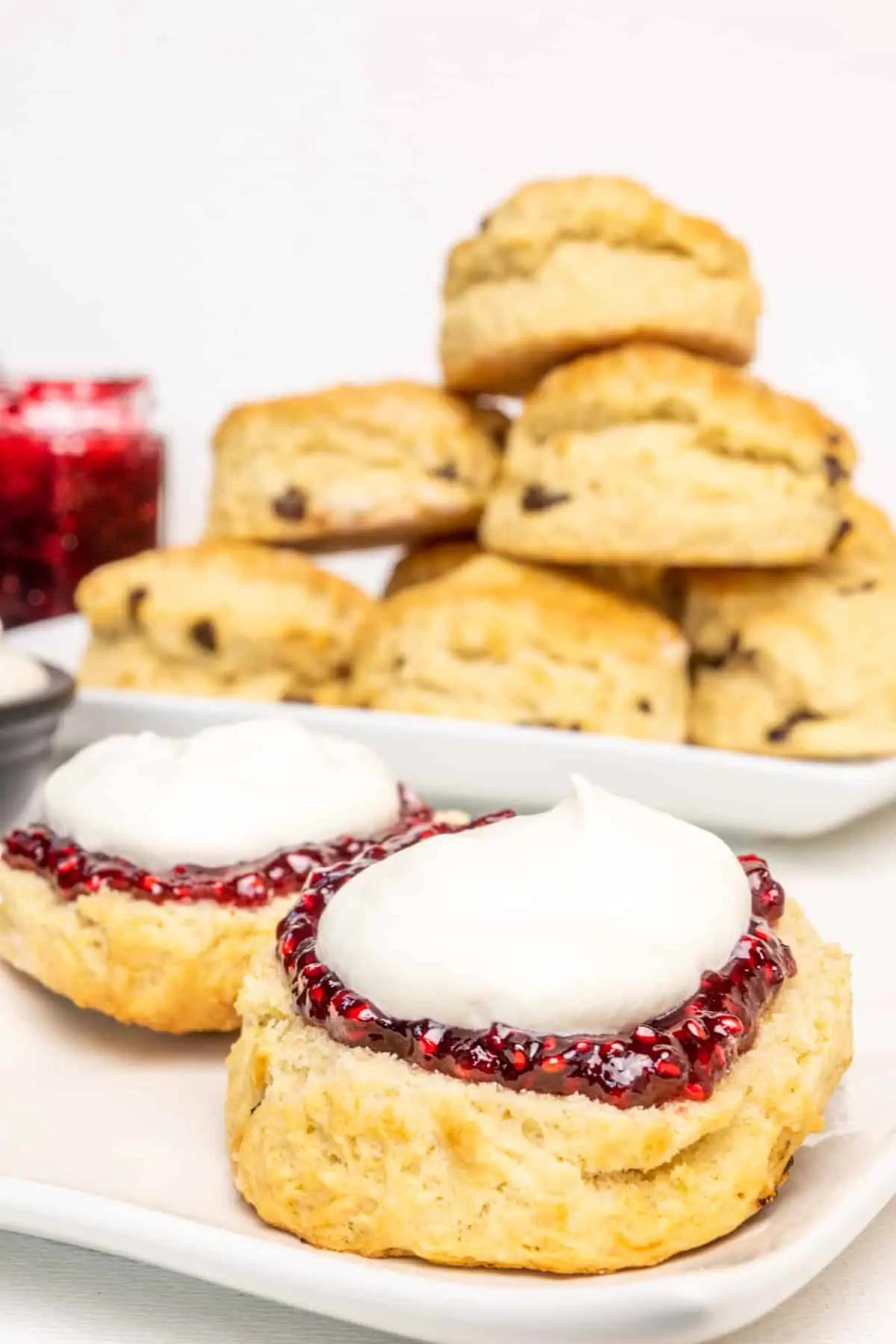 A scone halved and topped with raspberry jam and a dollop of whipped cream, in front of a stack of scones on a plate.